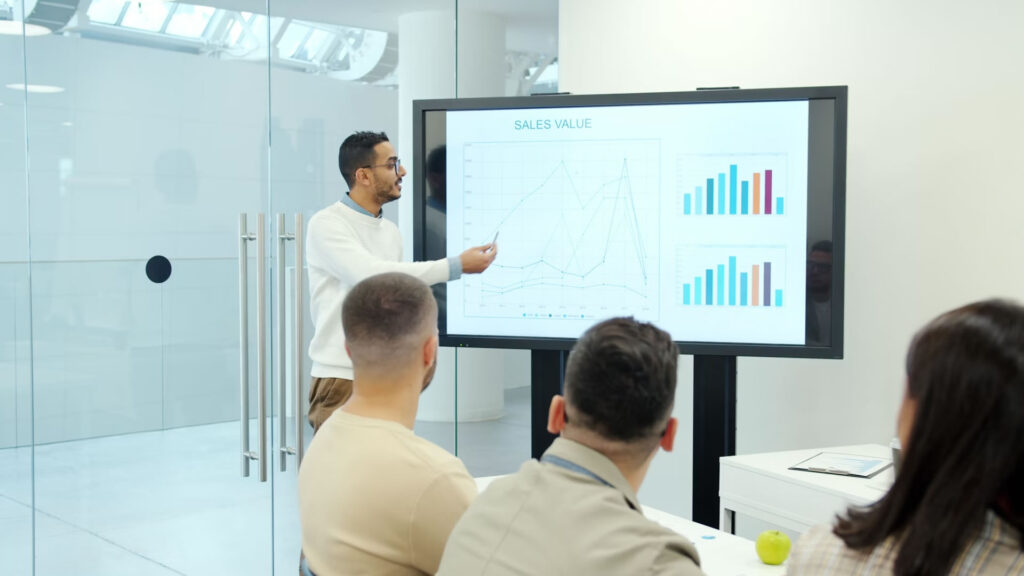 Business professional presenting sales charts and graphs on a screen to a team in a meeting room.