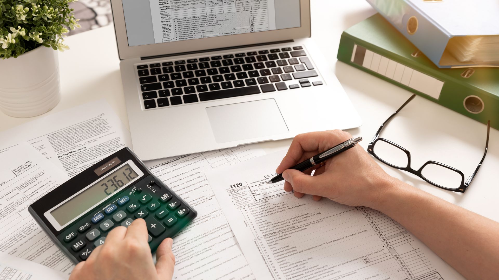 A business owner's hands using a calculator and filling out a form 1120 tax document on a desk covered in papers, with a laptop in the background.