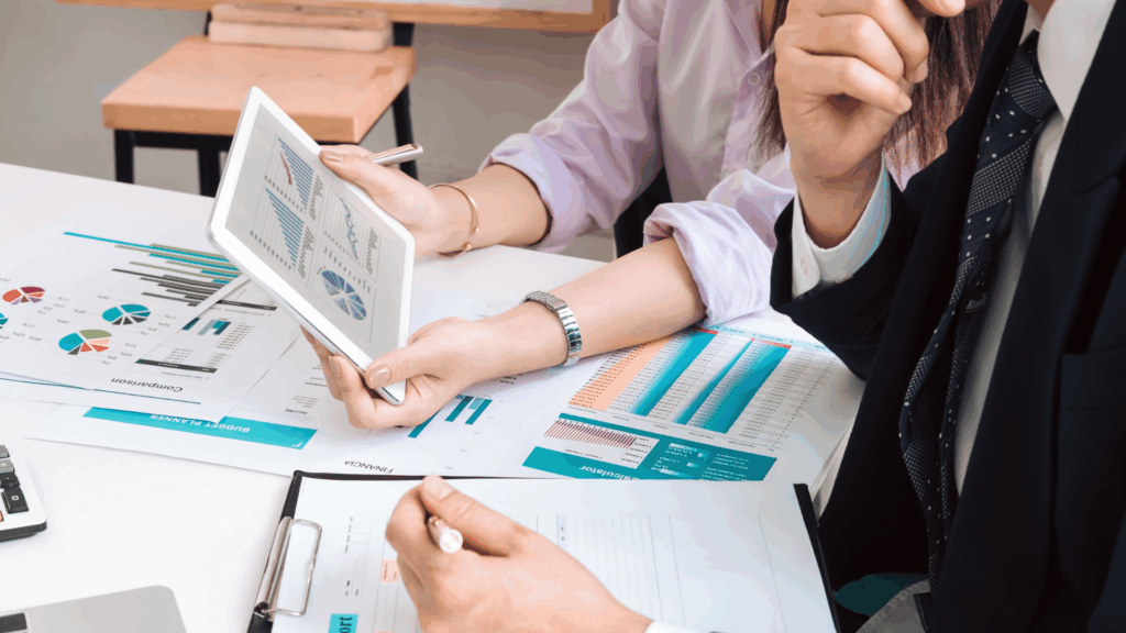 A business owner and a financial advisor reviewing charts and graphs on a tablet and paper documents to analyze business performance and cash flow.
