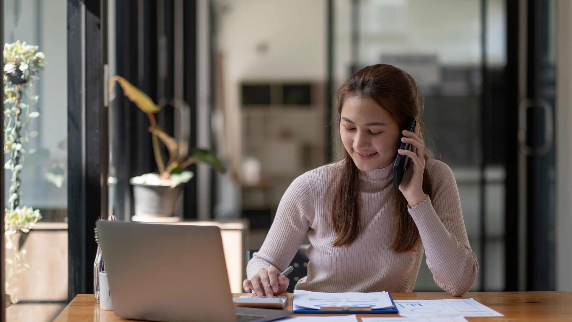 Businesswoman smiling while reviewing financial documents and talking on the phone at her desk