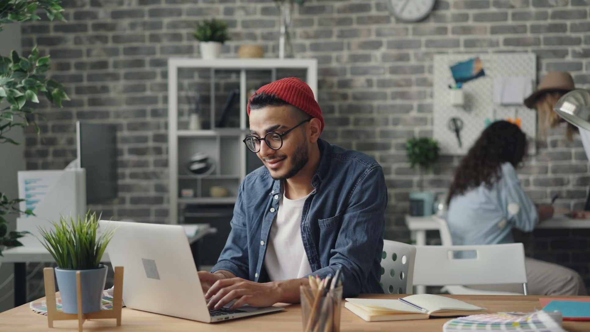 A young male entrepreneur, wearing glasses and a red beanie, smiling while working intently on a laptop at a modern office desk.