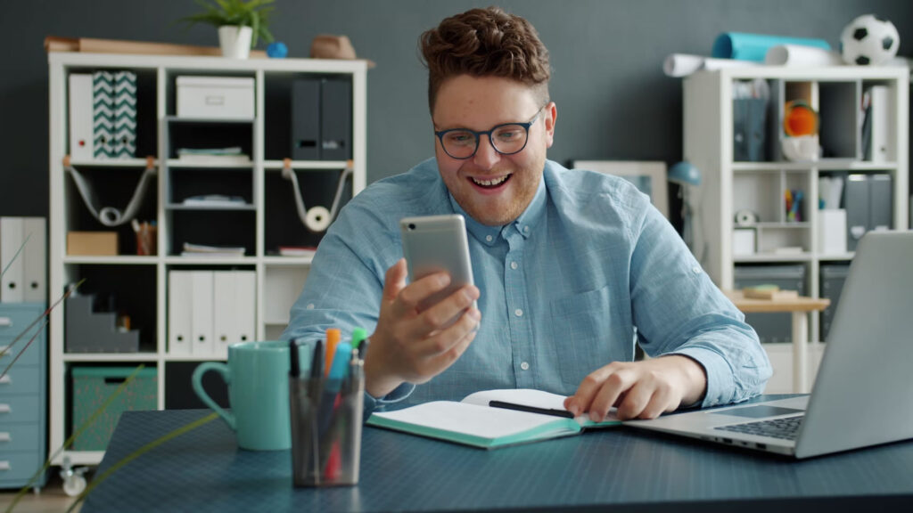 A smiling, glasses-wearing man in a blue shirt is actively managing his business finances using the Relay mobile app on his smartphone at his office desk.